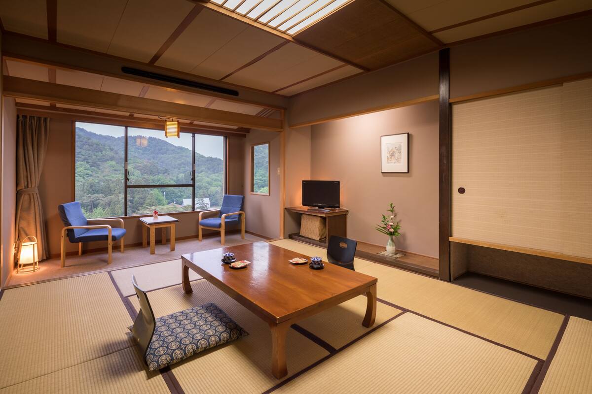 Ett traditionellt japanskt rum med tatamimattor på golvet, ett träbord, två blå stolar, en tv och utsikt över en skogbeklädd kulle.