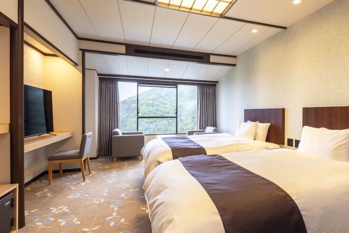 Habitación de hotel con dos camas, televisión, escritorio y ventana con vista a follaje.