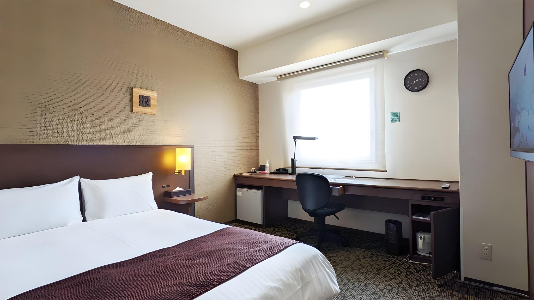 A hotel room with a large bed, a desk with a chair, a window, and a clock on the wall.