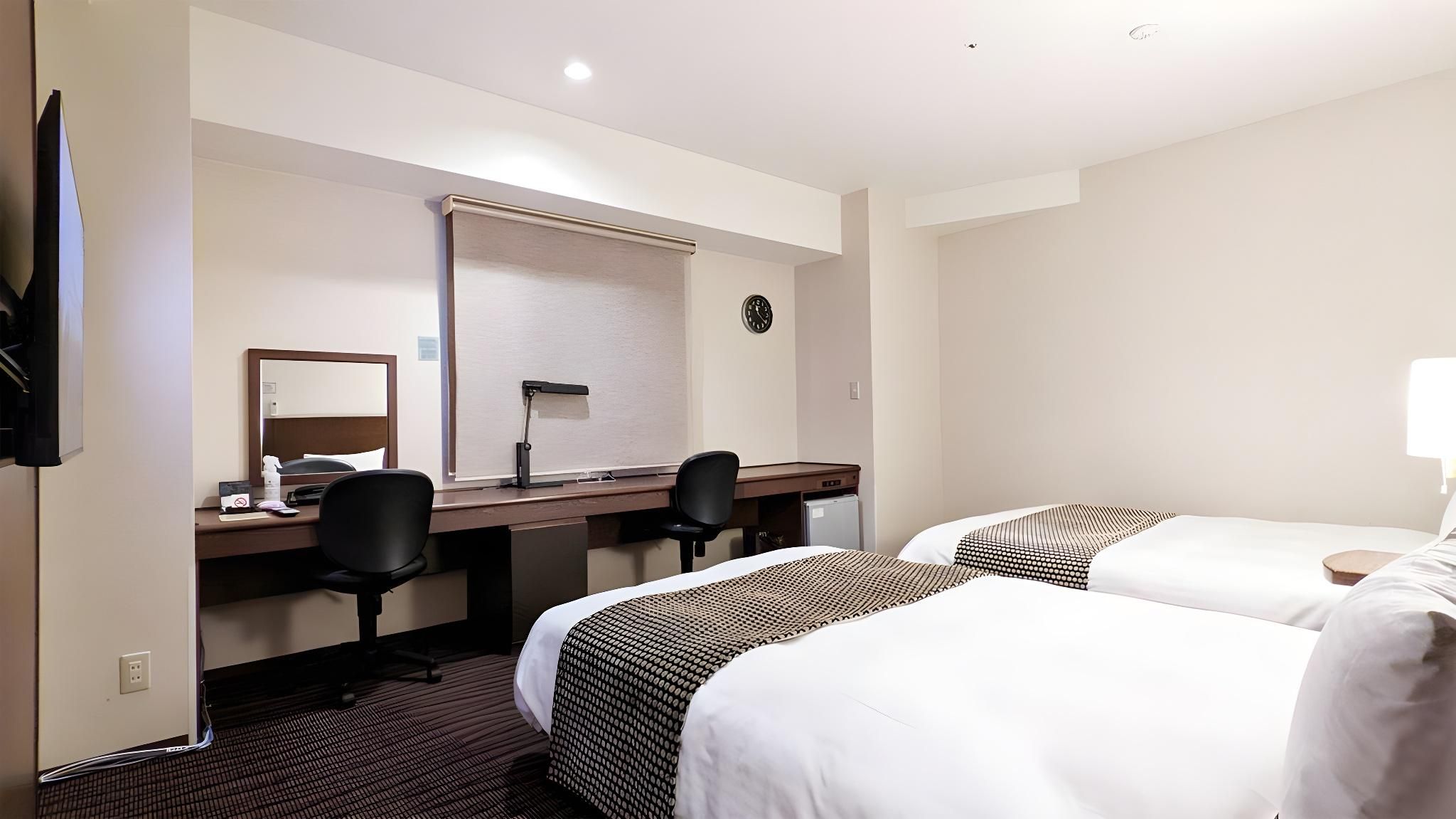 Habitación de hotel con dos camas, un escritorio con silla, un espejo, un reloj y un televisor.