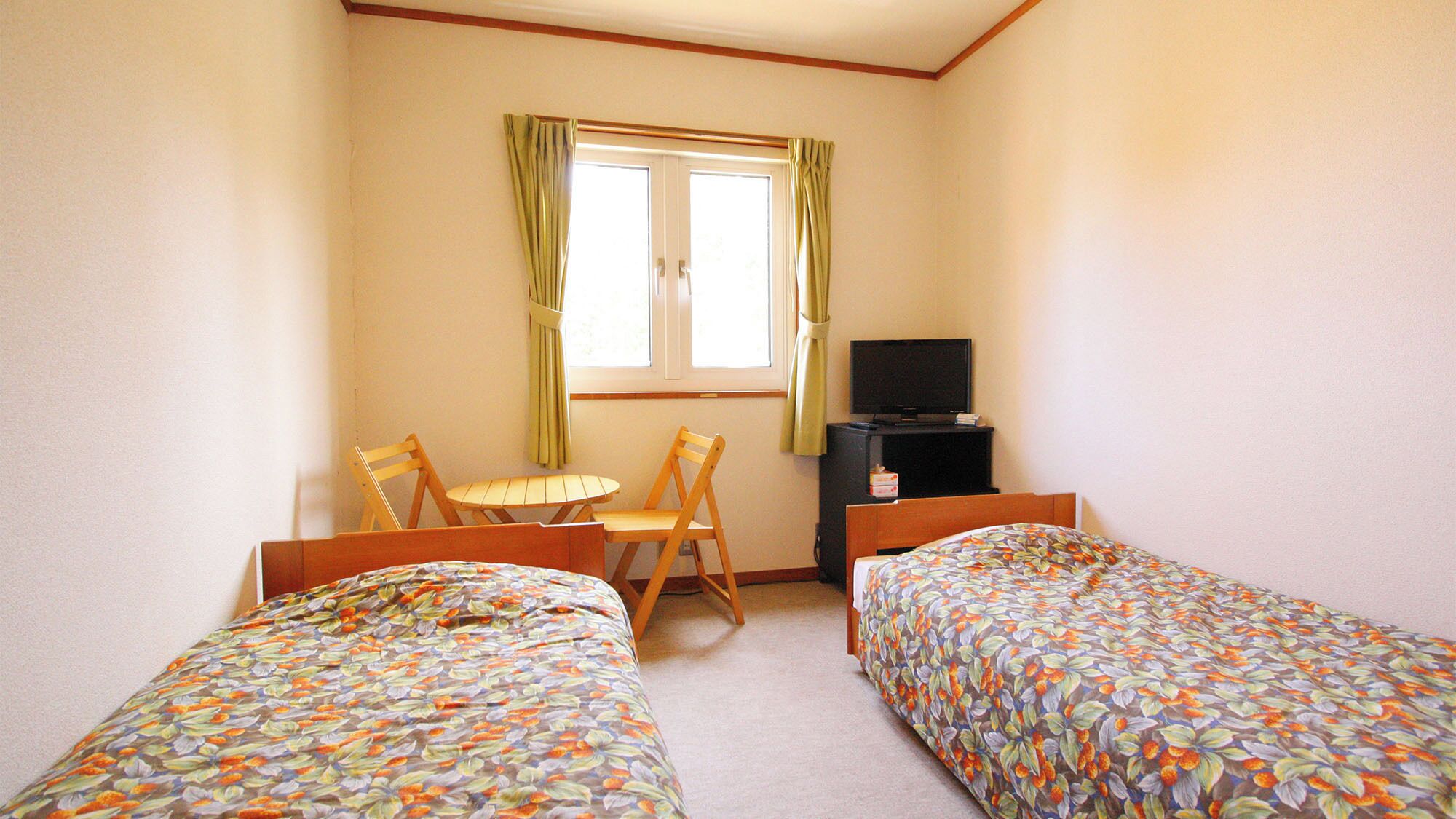 Una habitación con dos camas, una mesita, una silla, un televisor y una ventana con cortinas.