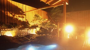 Vista nocturna de una estructura de madera con una zona iluminada, posiblemente una sauna o jacuzzi, rodeada de rocas y plantas.