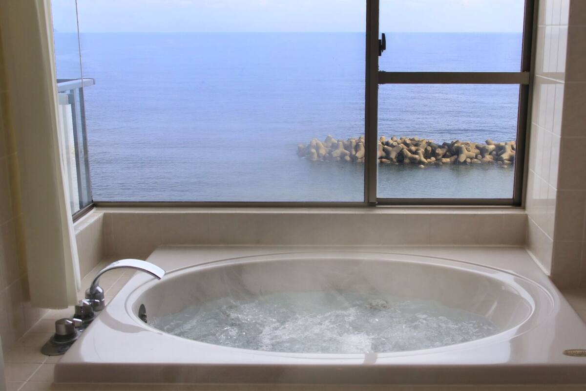 A bathroom with a bathtub, a window with a view of the sea, and a tiled floor.