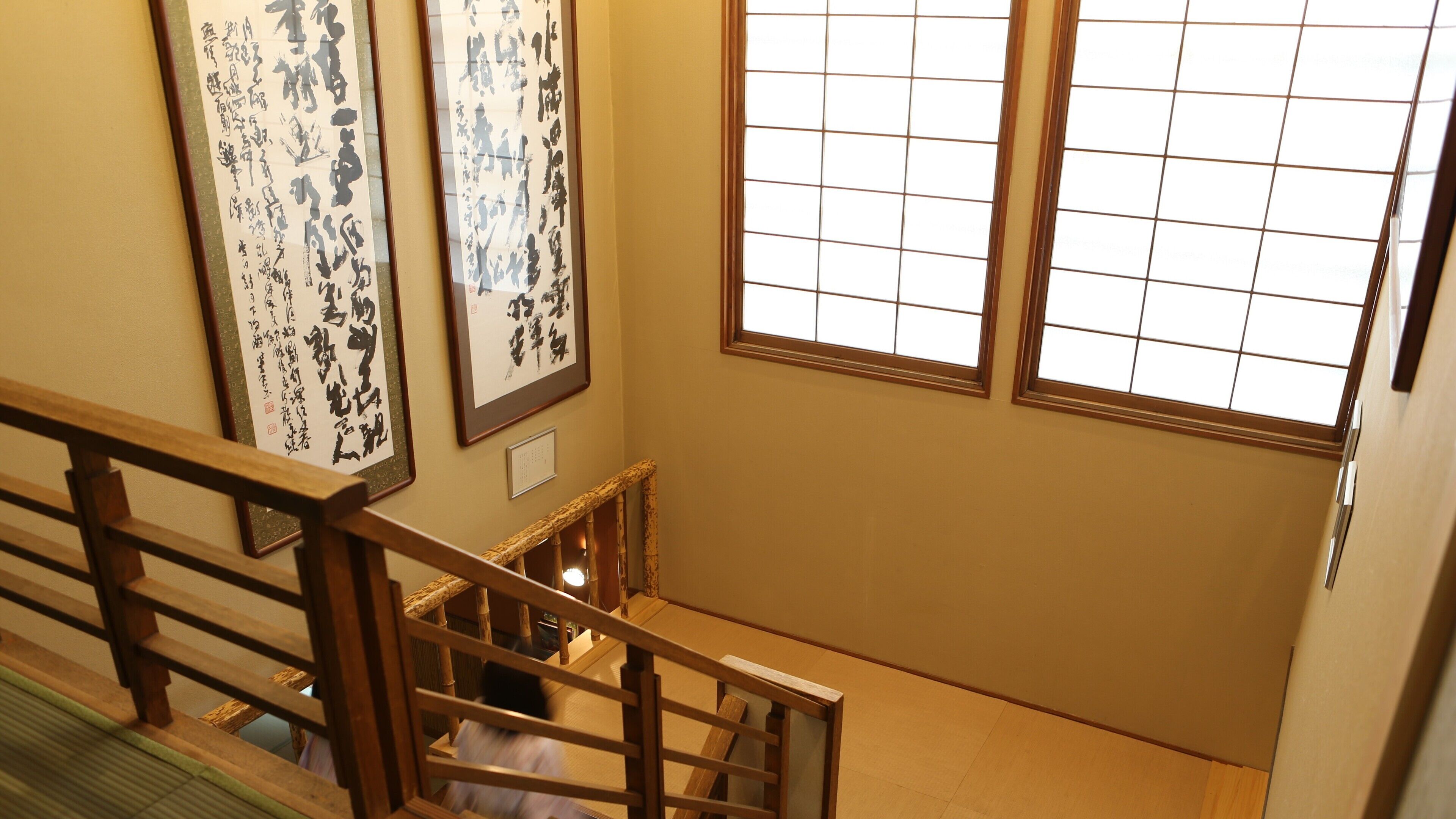 A staircase with wooden railings and a view of a room with traditional Japanese calligraphy art on the walls and a window with a grid pattern.
