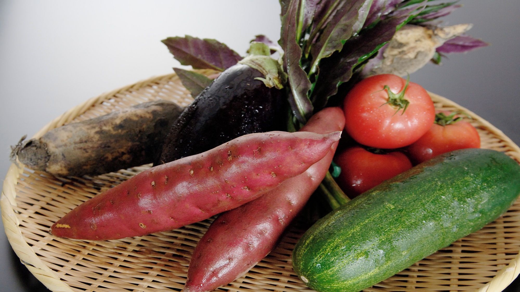 A variety of vegetables including sweet potatoes, a cucumber, and tomatoes on a woven basket.
