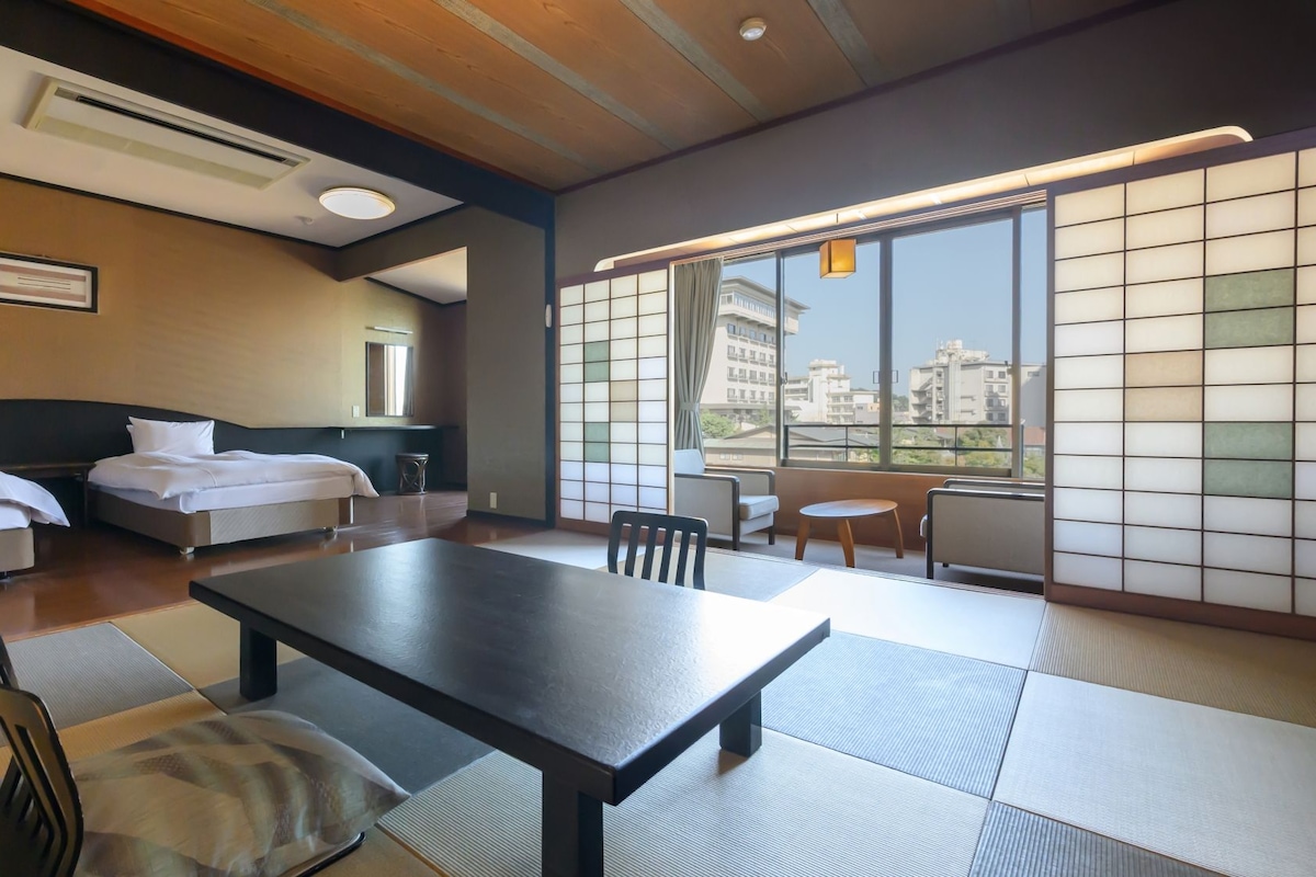 A hotel room with a bed, a table, chairs, a view of the city, and a traditional sliding door.