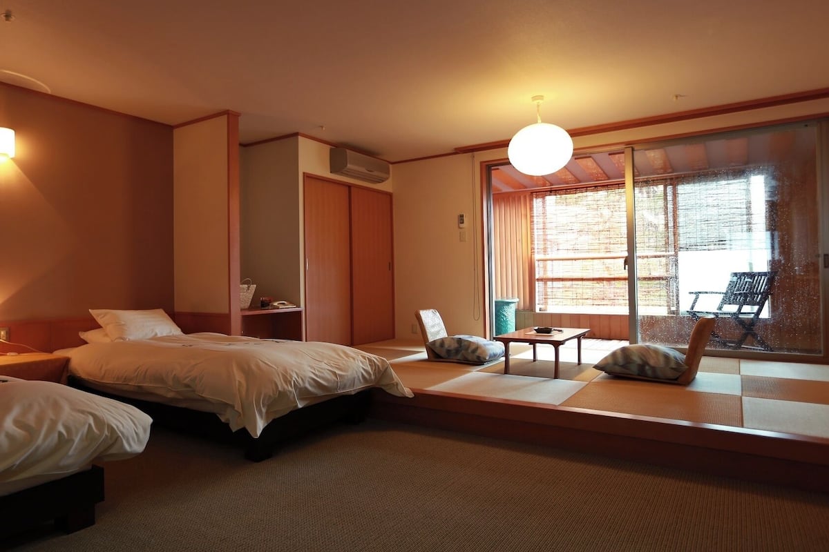 A hotel room with two beds, a small table, a chair, and a large window with a view.