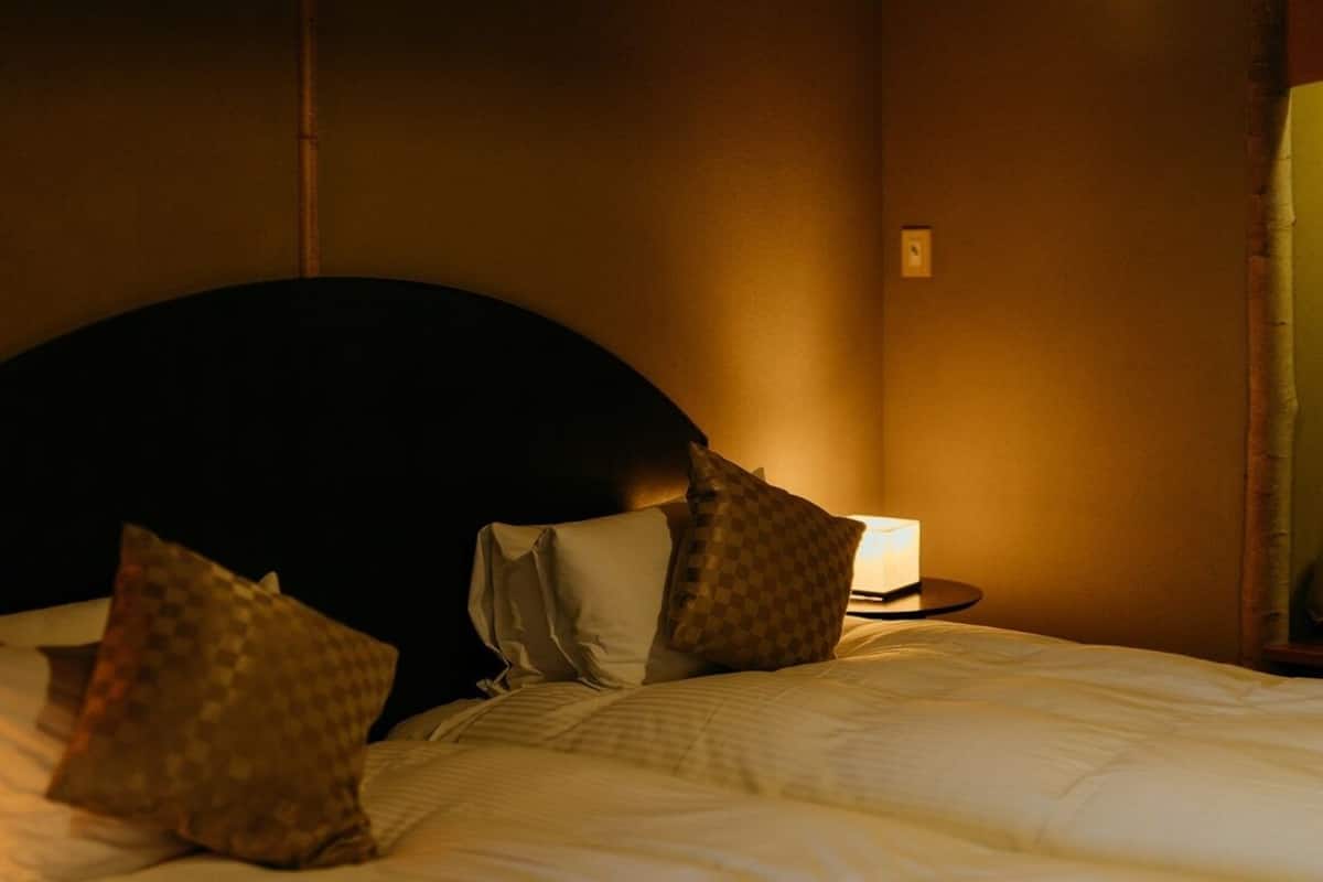 A hotel room with a bed, pillows, a bedside table with a lamp, and a wall-mounted light fixture.