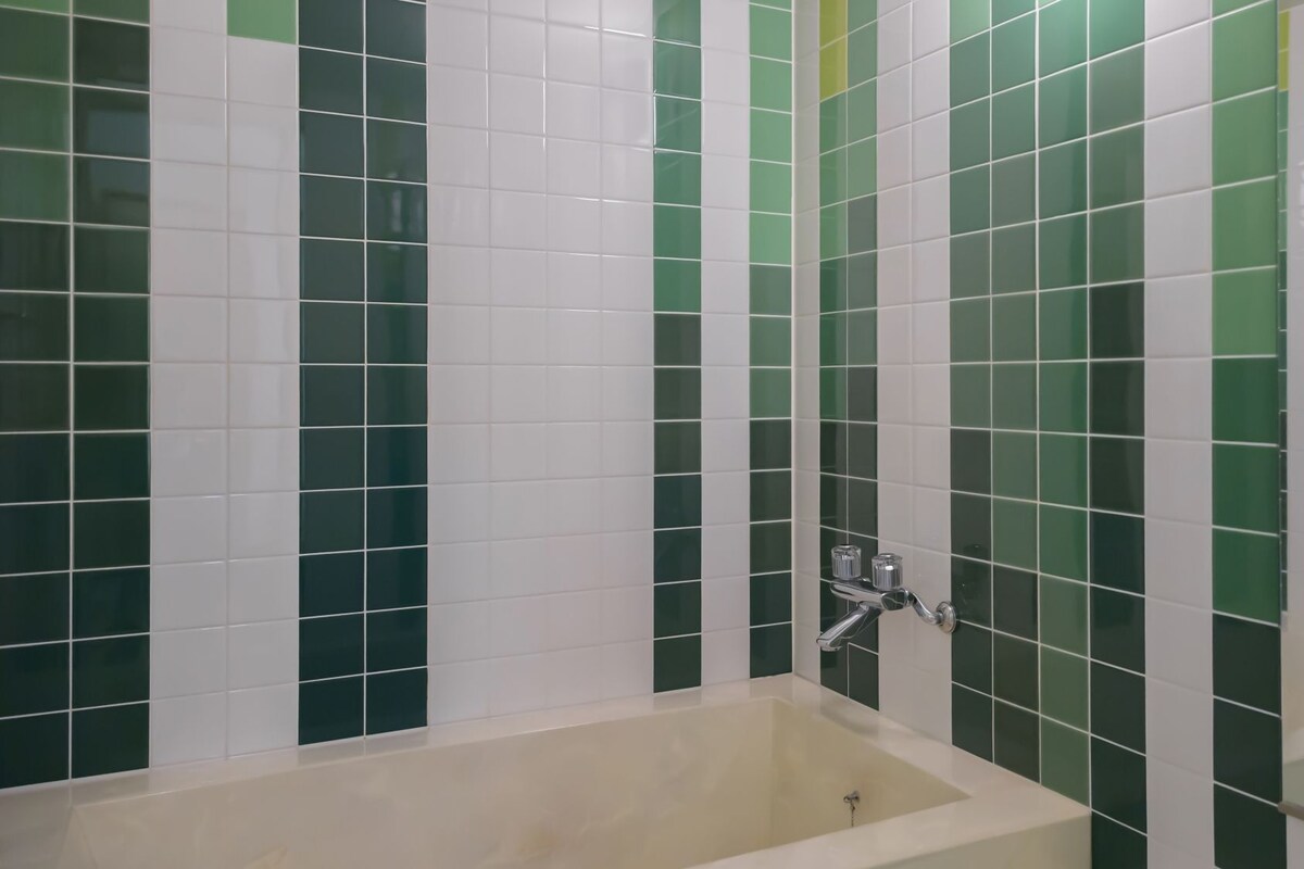 A bathroom with a bathtub, green and white tiled walls, and a chrome faucet.