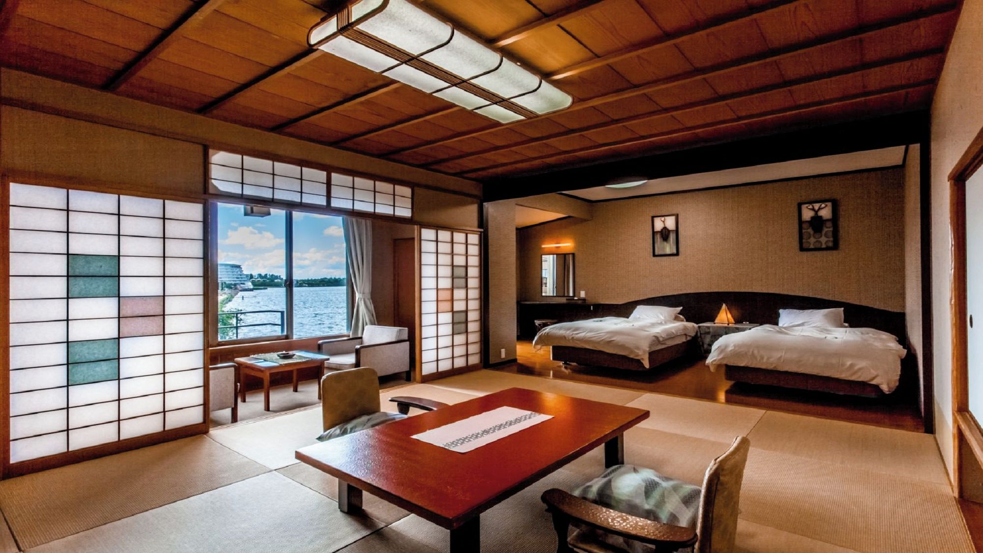 A traditional Japanese-style room with tatami flooring, a low wooden ceiling, and a view of a river. There are two beds, a table, and a chair.