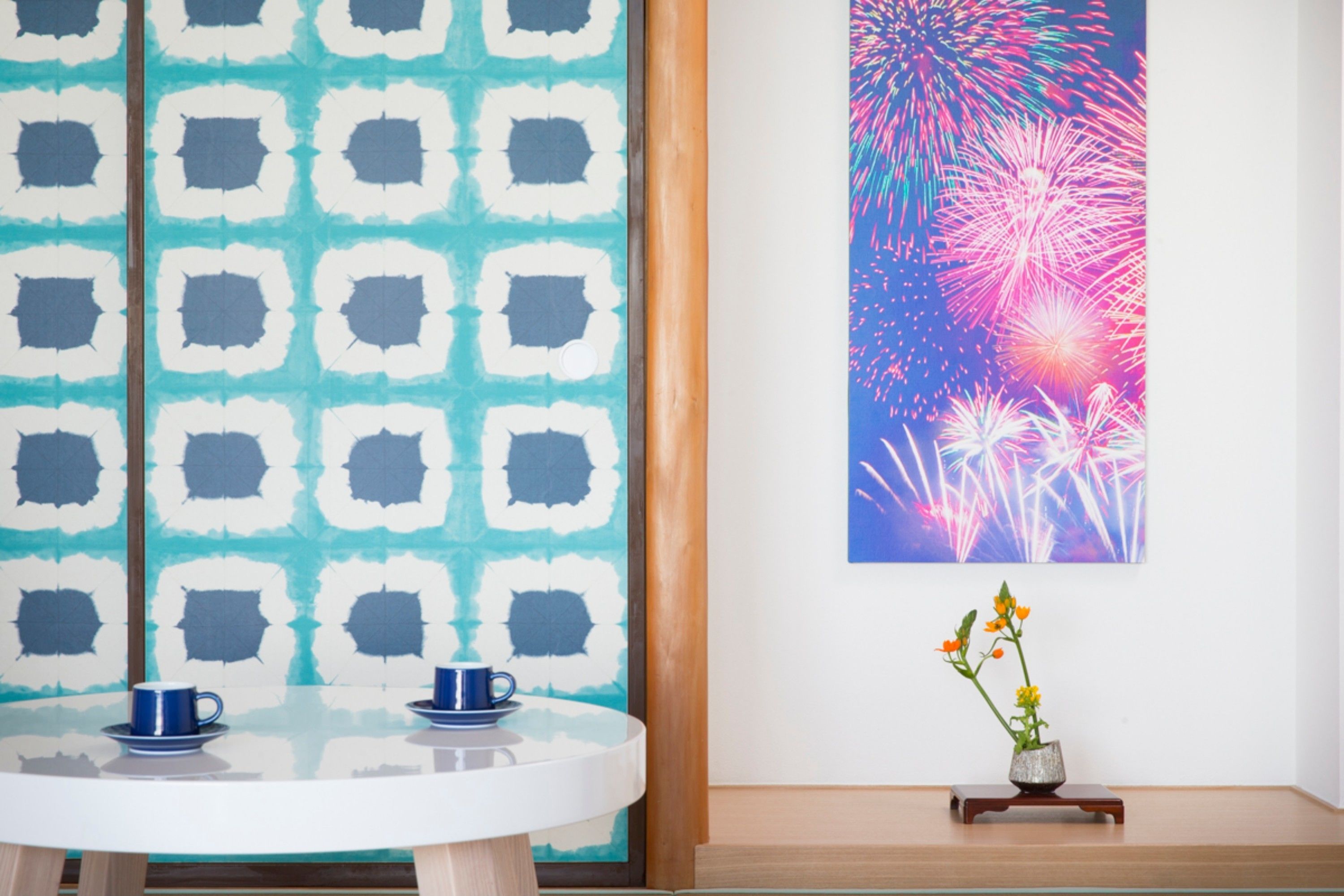 A room with a round table, two blue cups, a vase with flowers, and a colorful firework painting on the wall.