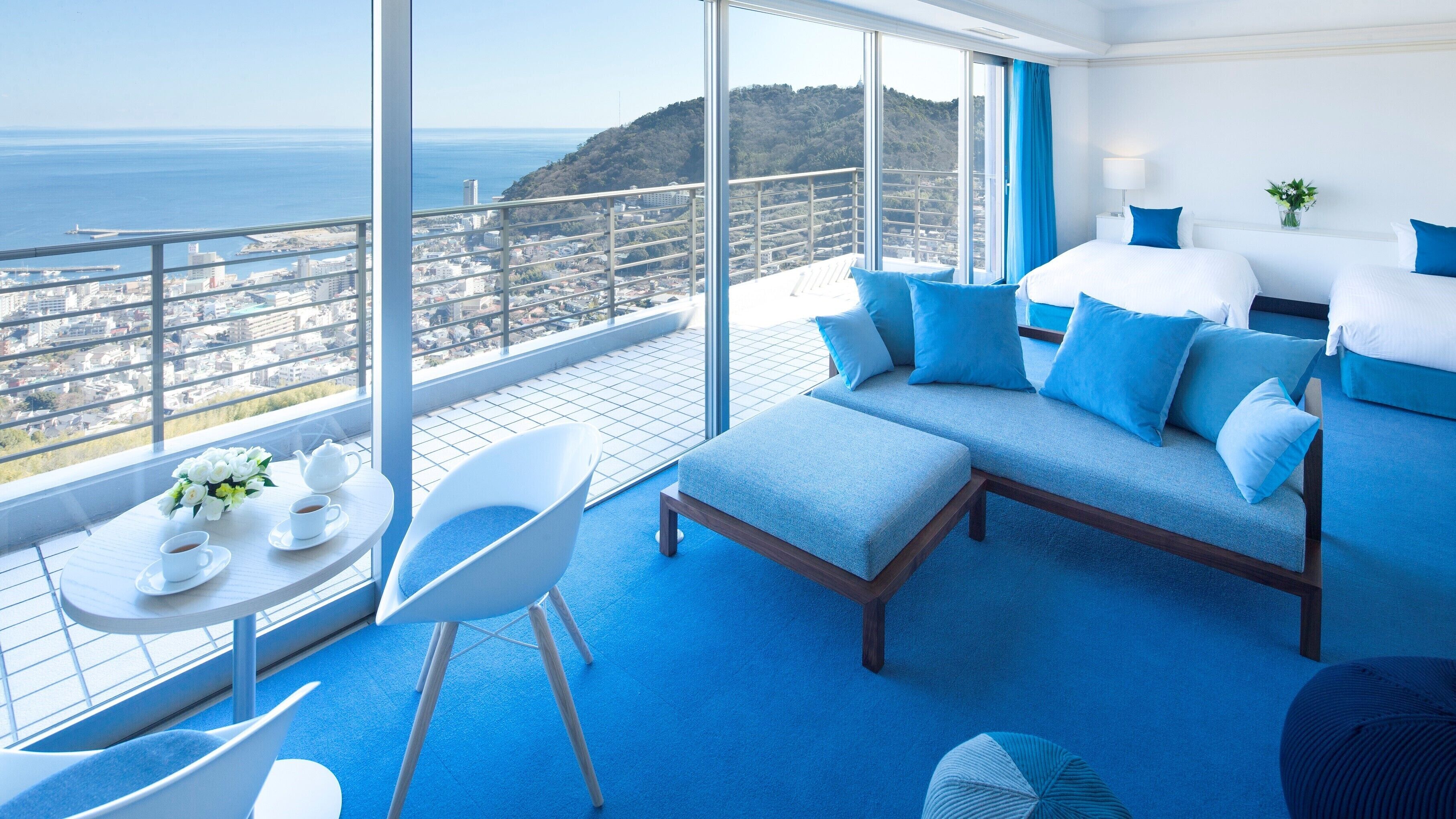 A balcony with a city view, a sofa, a chair, a table with a teapot and cups, and a bed with blue bedding.