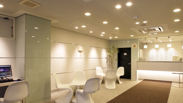 A modern reception area with a reception desk, several white chairs, a computer, and a plant. - Hotel Green Palace (Sendai)