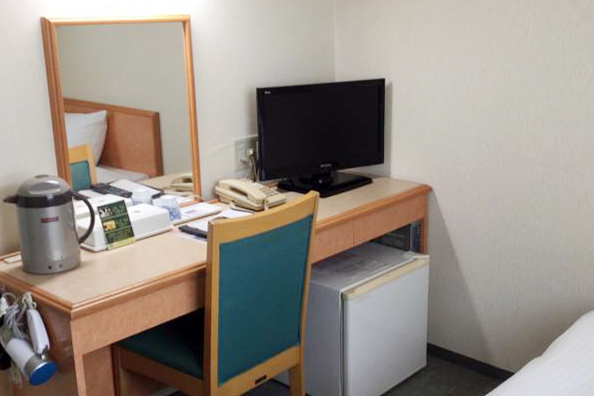 A hotel room with a wooden desk, a computer, a small refrigerator, a coffee maker, and a chair.