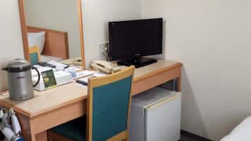 A hotel room with a wooden desk, a computer, a small refrigerator, a coffee maker, and a chair.