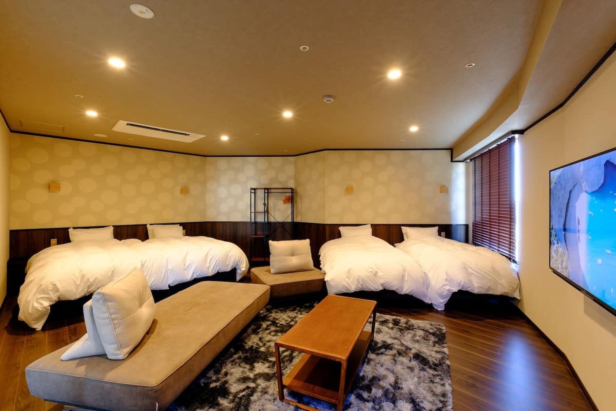 A hotel room with two beds, a sofa, a wooden bench, a rug, and a large window with blinds.