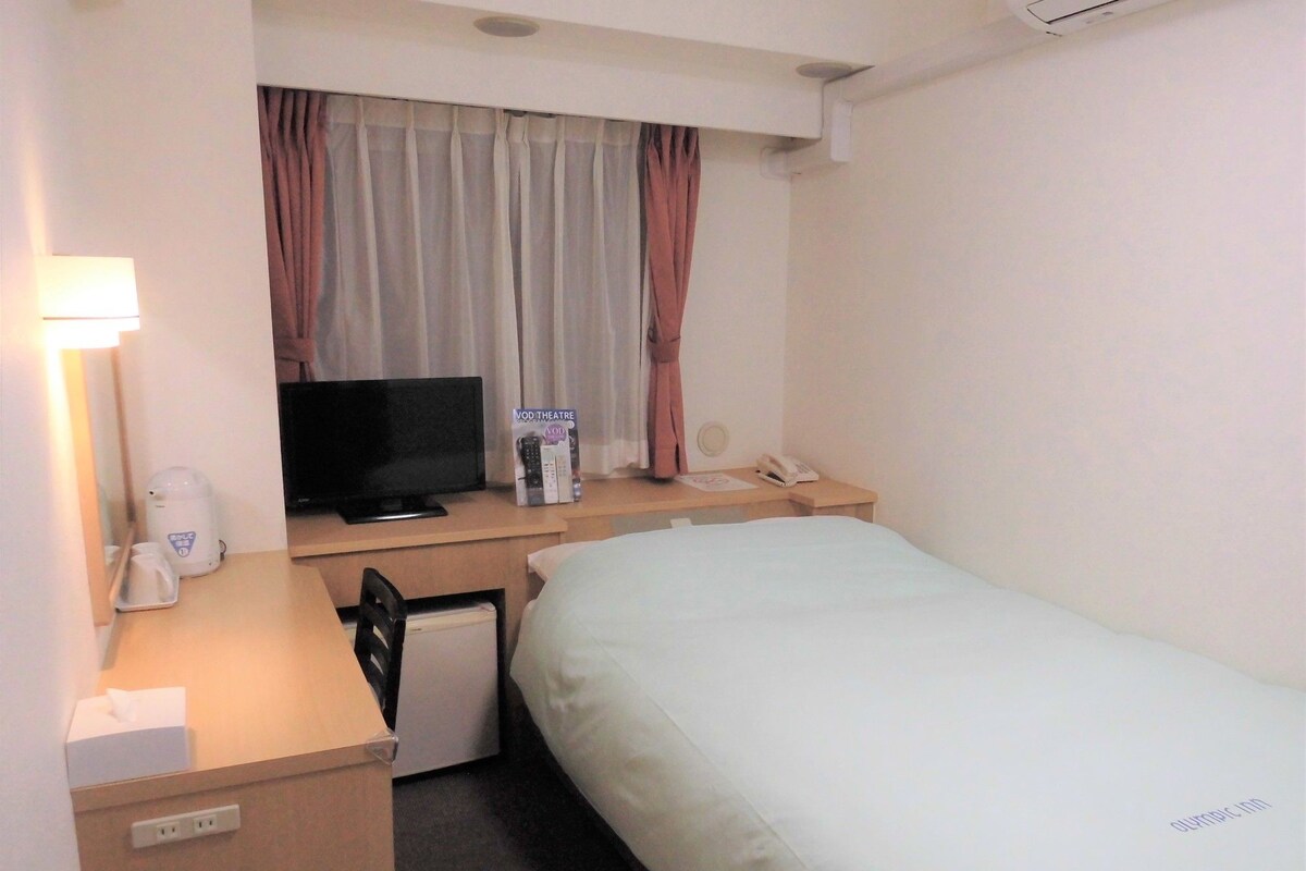 Une chambre d’hôtel comprenant un lit, un bureau avec une télévision, une machine à café, un téléphone et une fenêtre avec des rideaux.