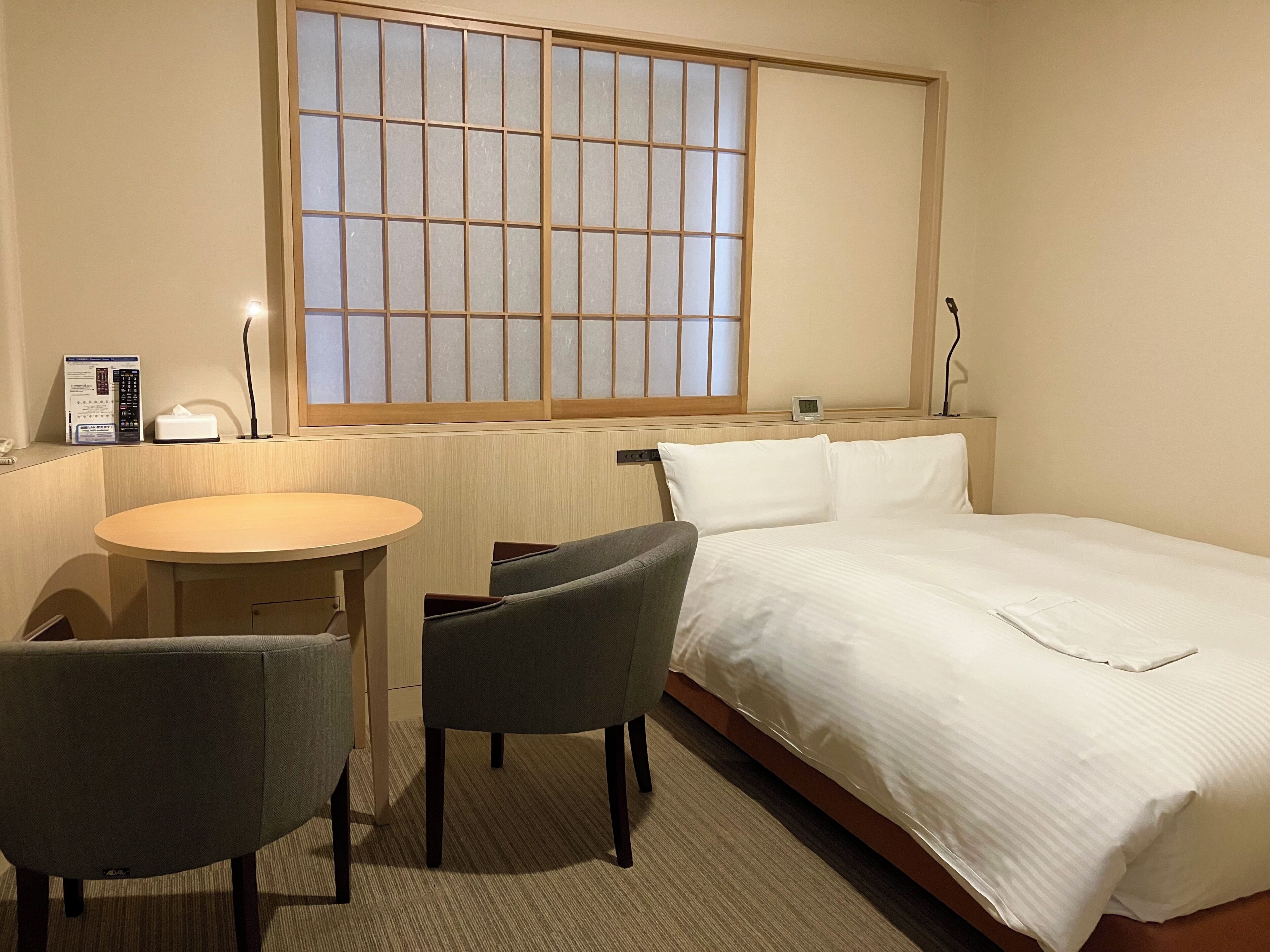Una habitación de hotel con cama, escritorio, dos sillas, ventana con diseño de cuadrícula y mesita de noche con lámpara.