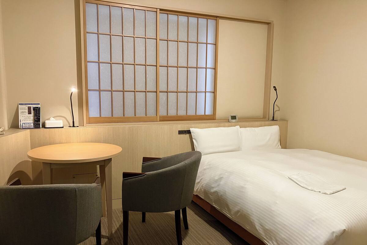 Una camera d'albergo con un letto, una scrivania, due sedie, una finestra con un motivo a griglia e un comodino con una lampada.