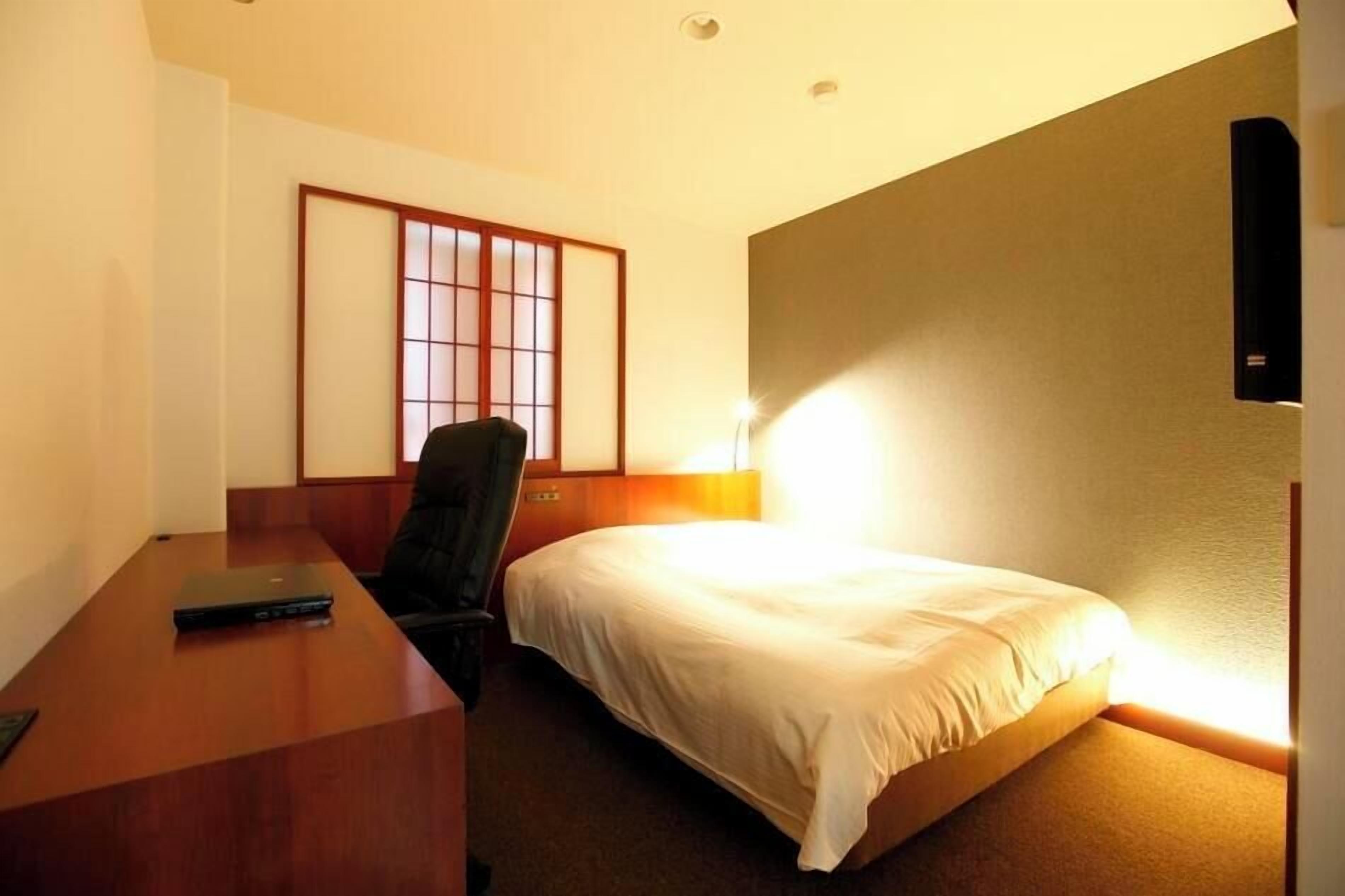 A hotel room with a bed, a desk, a chair, a wall-mounted TV, and a traditional Japanese shoji screen.