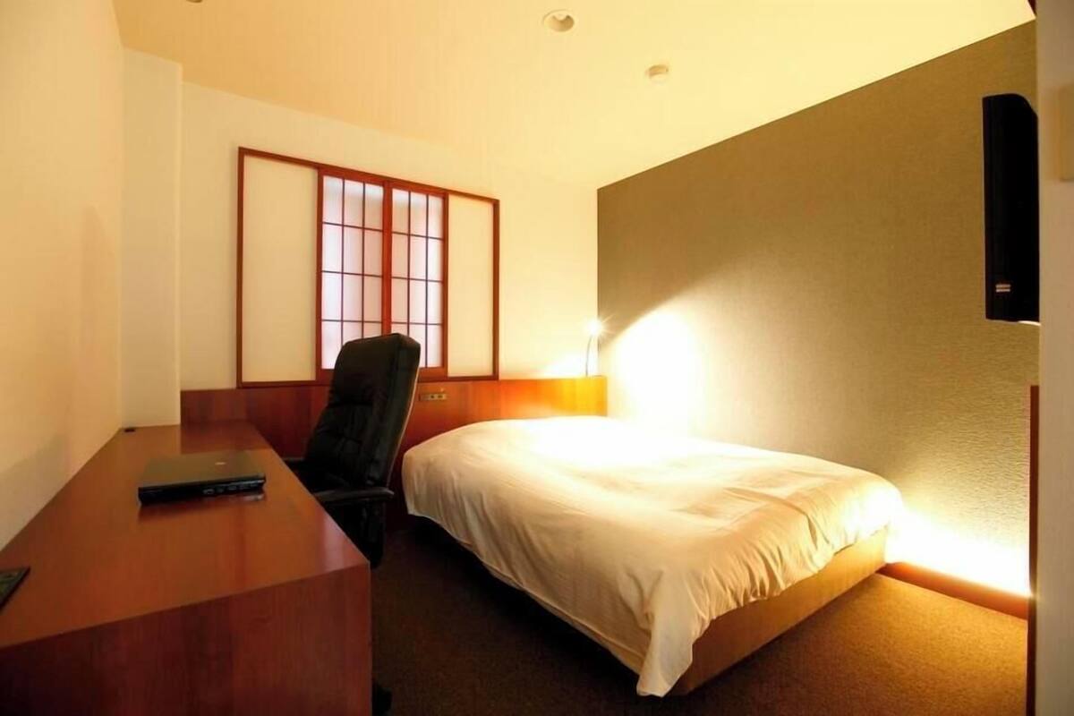 Una camera d'albergo con un letto, una scrivania, una sedia, una televisione a muro e un tradizionale schermo giapponese shoji.