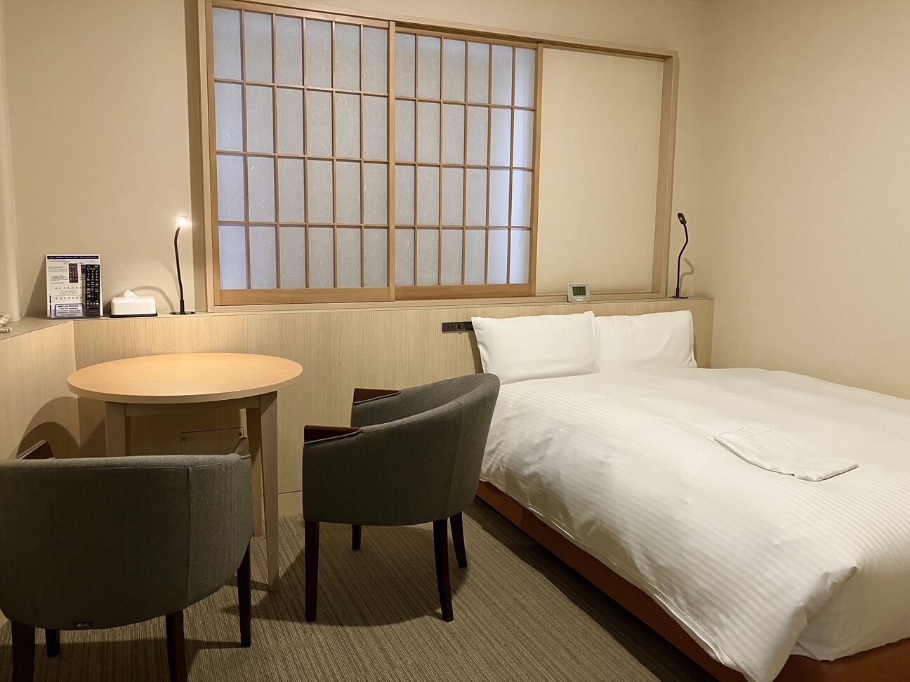 A hotel room with a bed, a small table, two chairs, a window with a view, and a bedside table with a lamp.