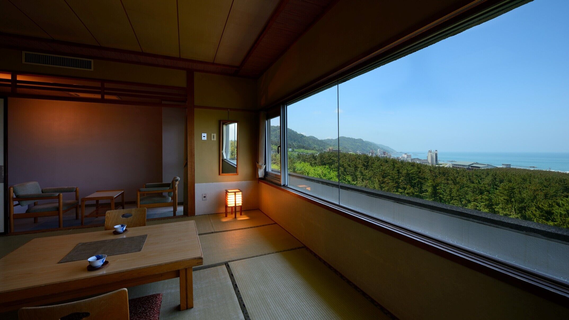 A traditional Japanese room with tatami flooring, a low table, and chairs. A large window offers a view of a coastal landscape.