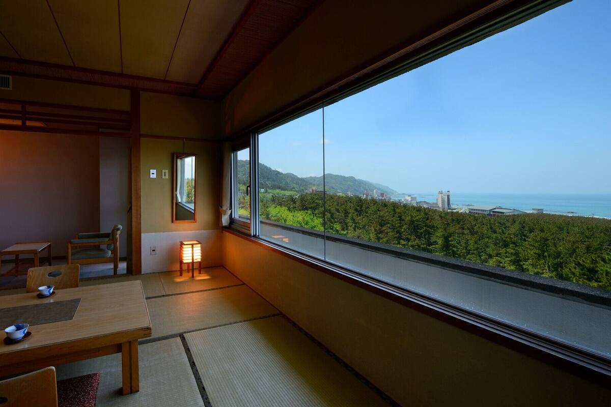 A traditional Japanese room with tatami flooring, a low table, and chairs. A large window offers a view of a coastal landscape.