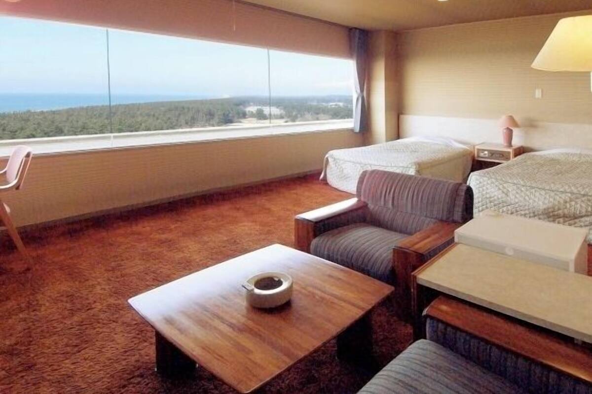 A hotel room with a large window, a bed, a sofa, a small table, and a chair.