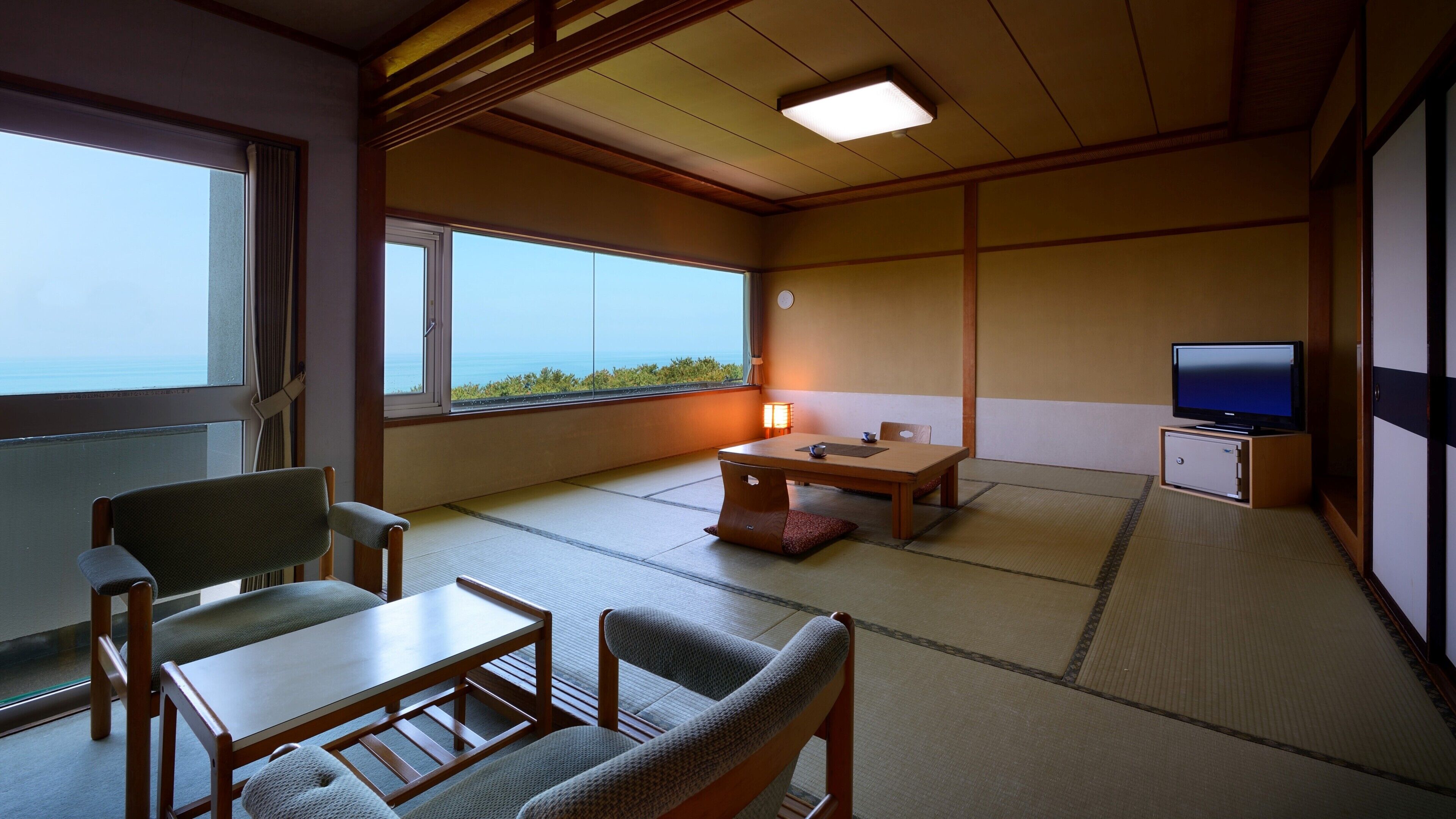 A traditional Japanese room with tatami flooring, a low table, chairs, a television, and a view of the sea.
