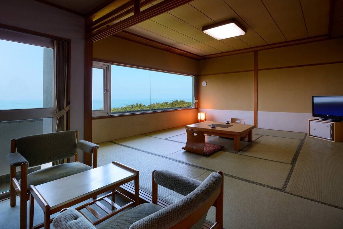 A traditional Japanese room with tatami flooring, a low table, chairs, a television, and a view of the sea.