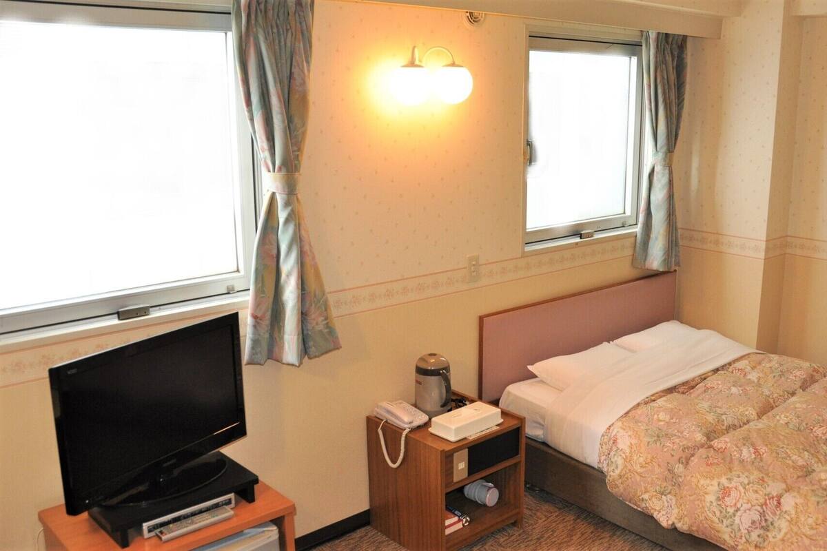 Habitación de hotel con cama, televisor, mesita de noche con teléfono y tetera, y cortinas en las ventanas.