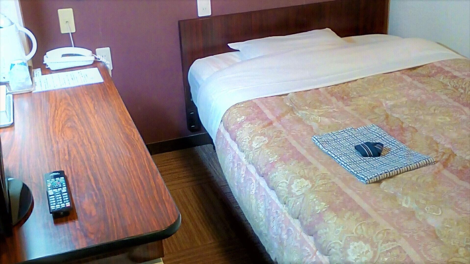 A hotel room with a wooden desk, a bed with a patterned bedspread, a remote control on the desk, and a wall socket.
