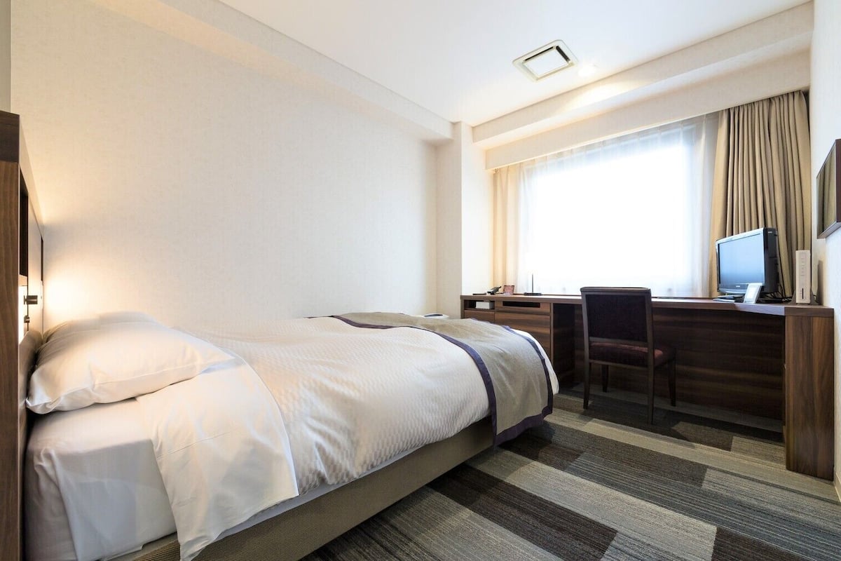 Una camera d'albergo con un letto, una scrivania con un computer e una finestra con tende.