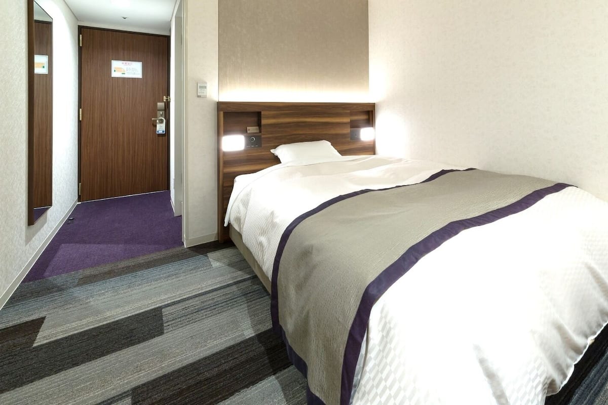 Una camera d'albergo con un letto, un comodino, una lampada e un pavimento rivestito di moquette.