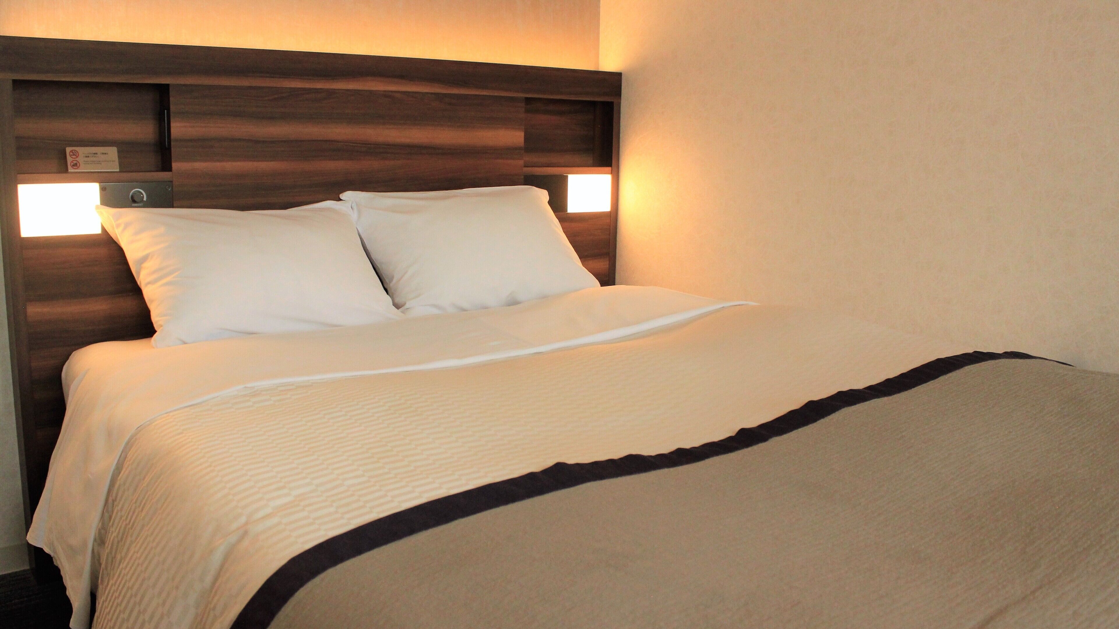 Hotelkamer met houten hoofdeinde, twee witte kussens, een beige sprei en een zwarte rand aan het voeteneinde van het bed.