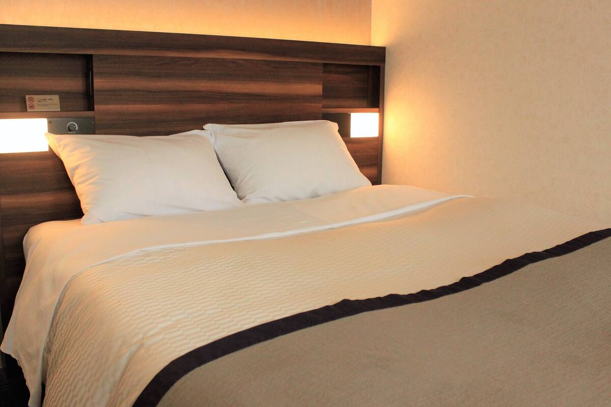 Camera d'albergo con testiera in legno, due cuscini bianchi, copripiumino beige e finitura nera alla base del letto.