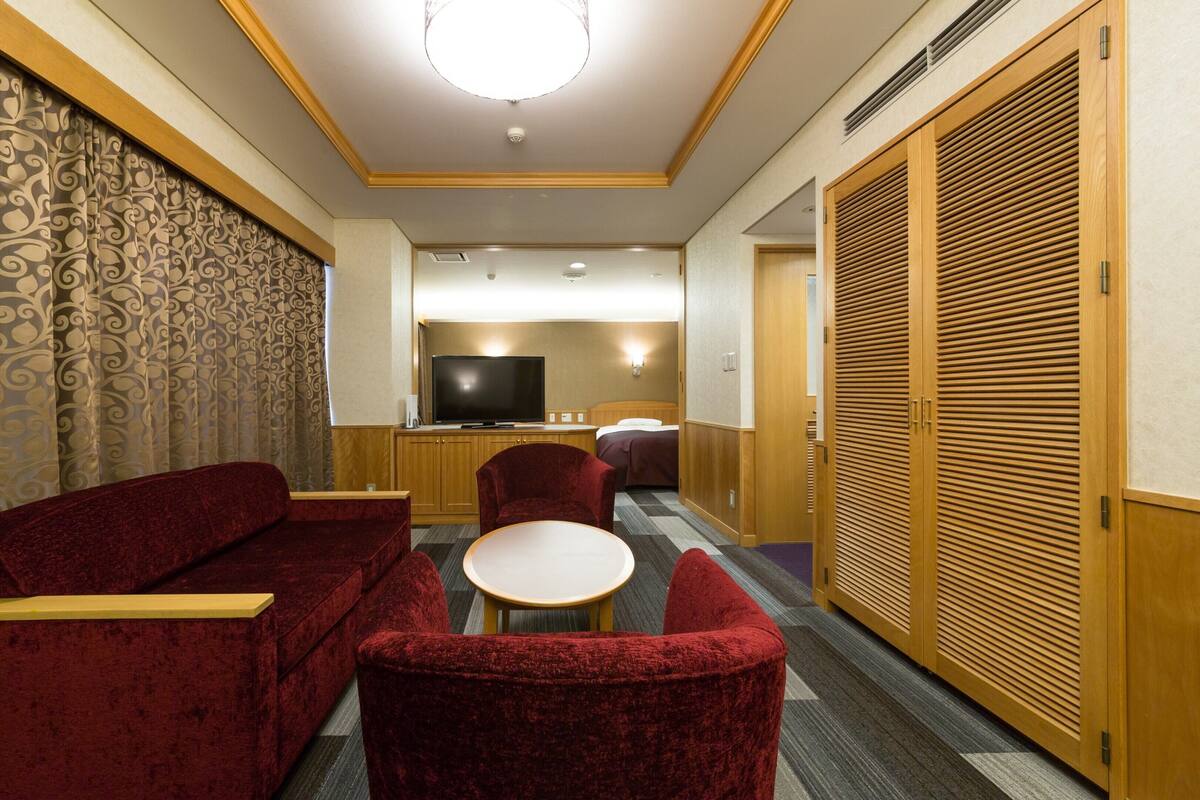 Camera d'albergo con divano, poltrona e un tavolino. Si vede una televisione, un letto e un'armadio.