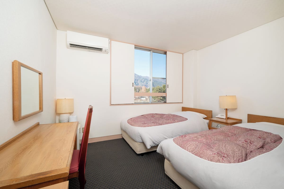 Een hotelkamer met twee bedden, een houten bureau, een stoel, een raam met uitzicht op bergen en een airconditioner.