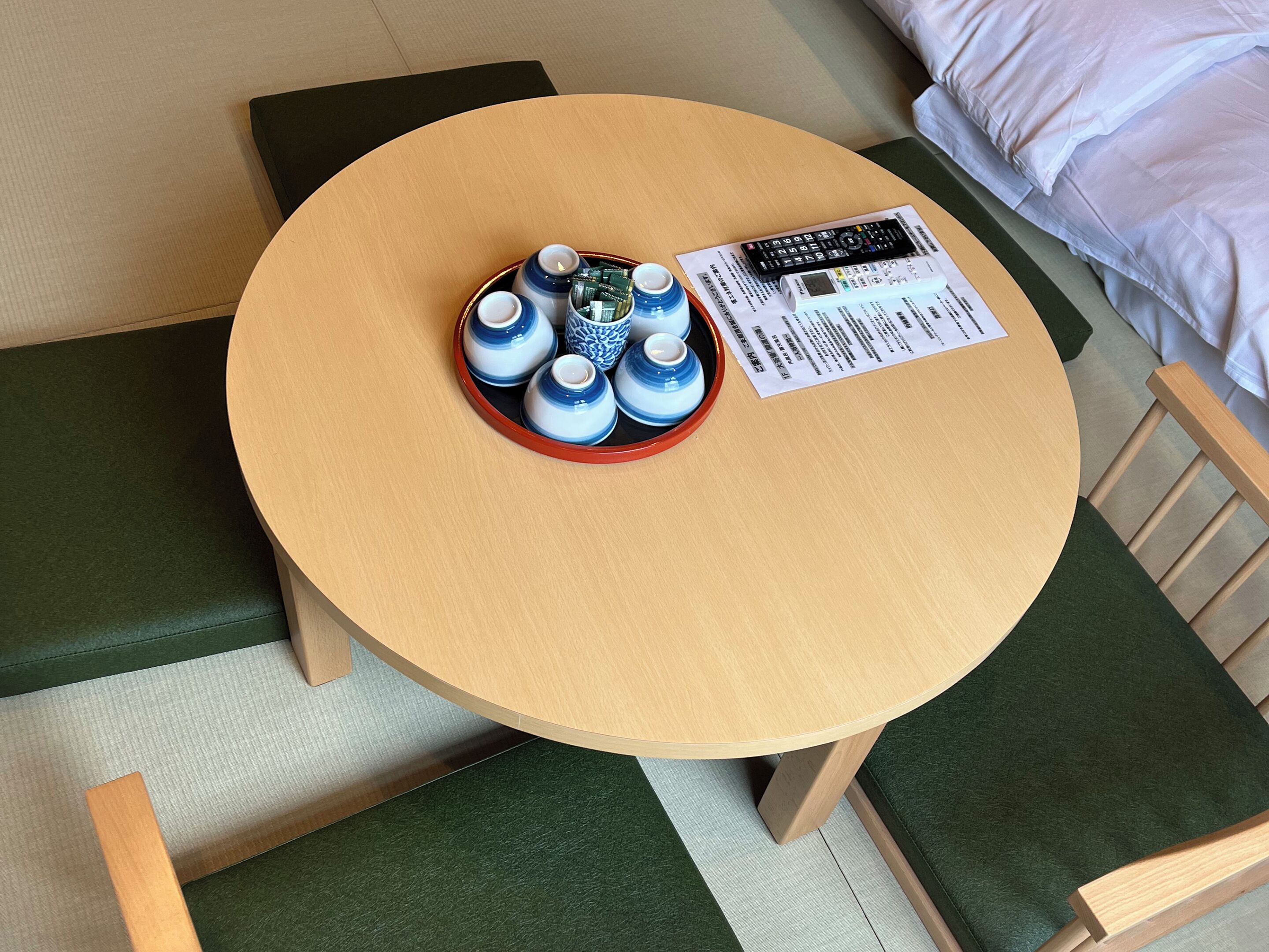 Une table ronde en bois, sur laquelle se trouve un plateau avec des théières bleues et blanches, une télécommande et un menu, le tout posé sur un banc vert rembourré.