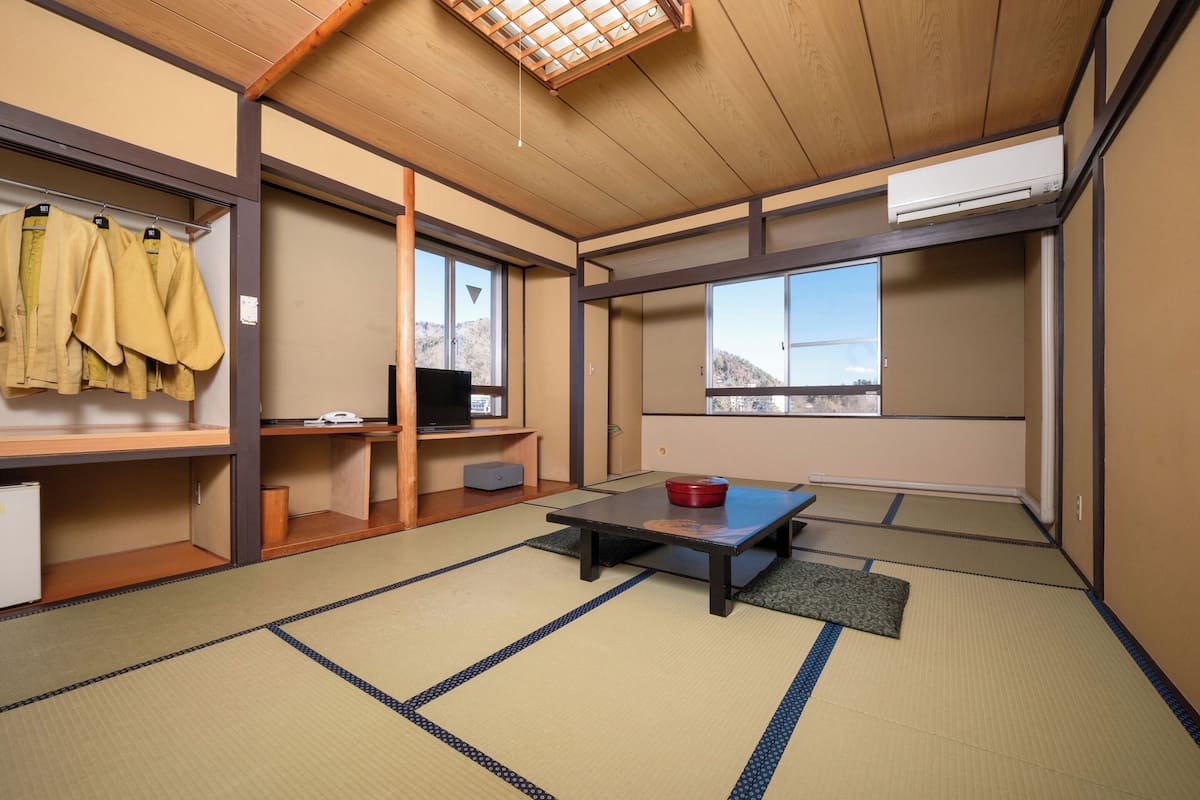Een traditionele Japanse kamer met tatami vloerbedekking, een lage tafel met een rode kom, een televisie op een plank en een kledingkast met hangende kleding.