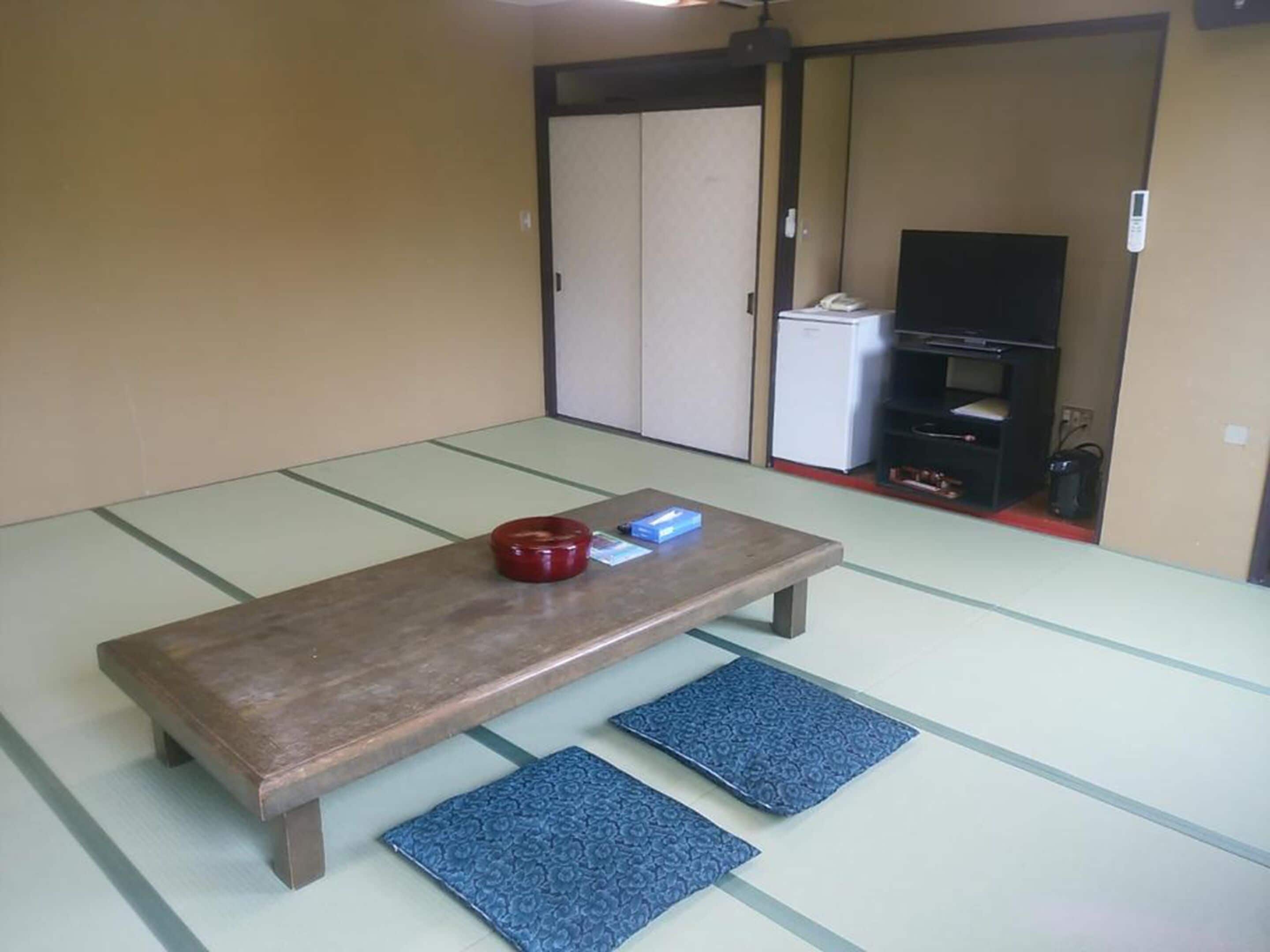 A tatami mat room with a wooden table, two blue cushions, a red bowl, and a television in the background.