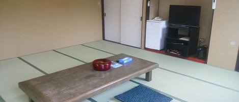 A tatami mat room with a wooden table, two blue cushions, a red bowl, and a television in the background.