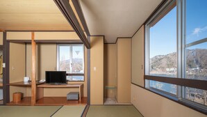 Ein traditioneller japanischer Raum mit Tatami-Boden, einem Schreibtisch mit Computer und einem großen Fenster mit Blick auf eine gebirgige Landschaft.