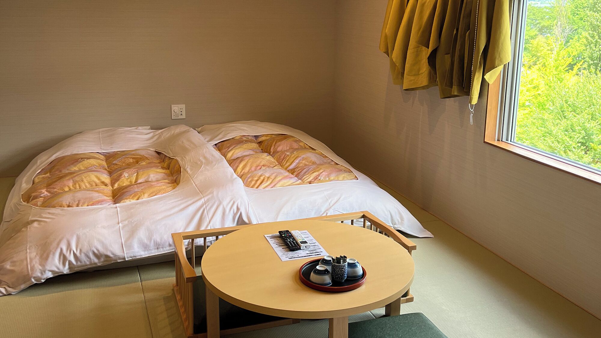 Een enkel bed met een deken met witte en gele strepen, een klein houten tafeltje met een afstandsbediening en een dienblad met kopjes, en een raam met gordijnen.