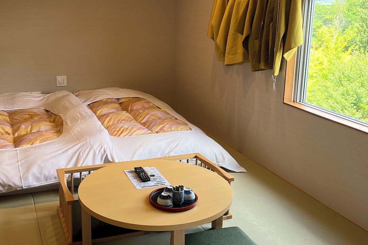 A single bed with a white and yellow striped duvet, a small wooden table with a remote control and a tray with cups, and a window with curtains.