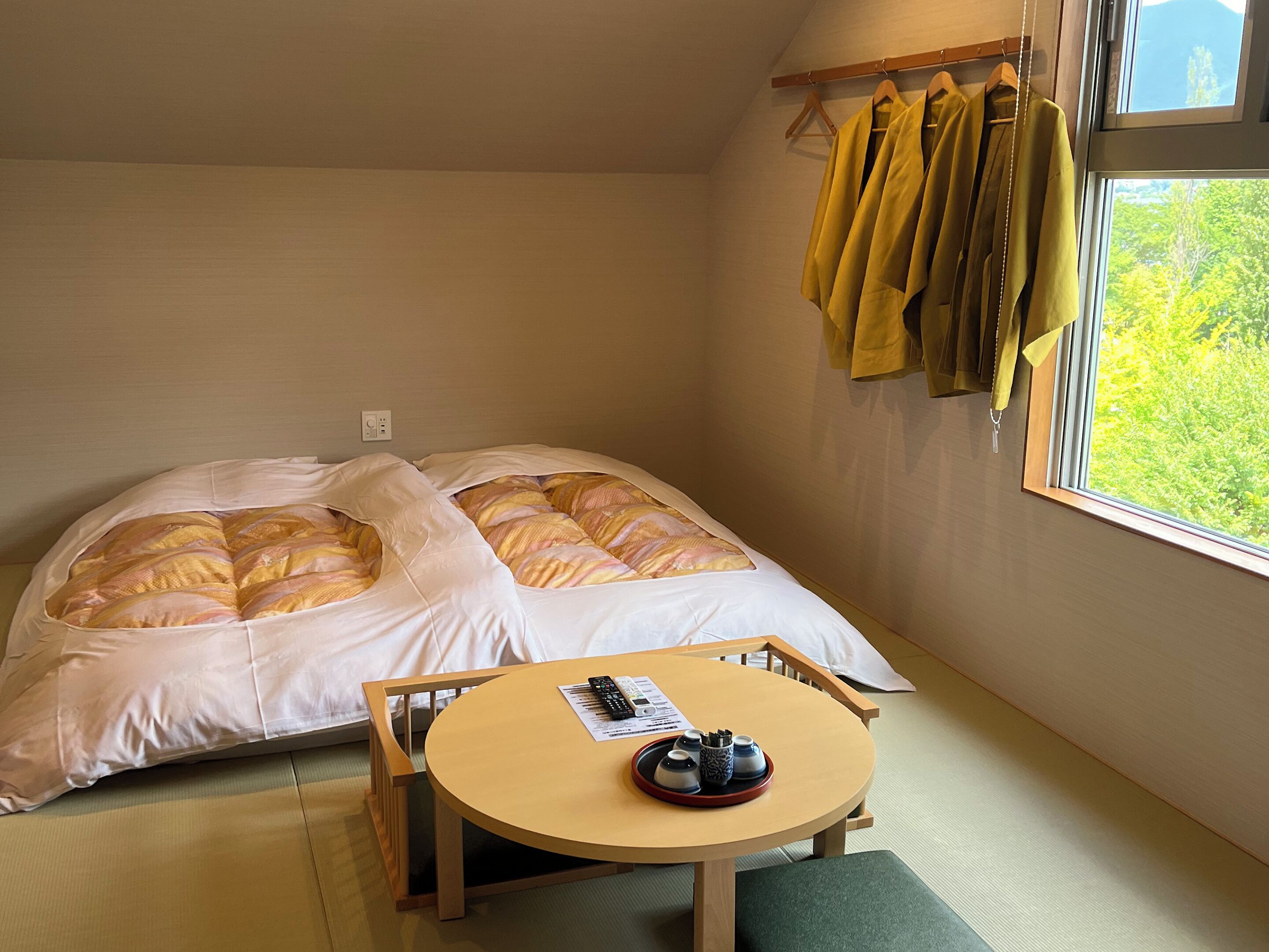 A room with a bed, a table with a remote control, and a window with a view of trees.