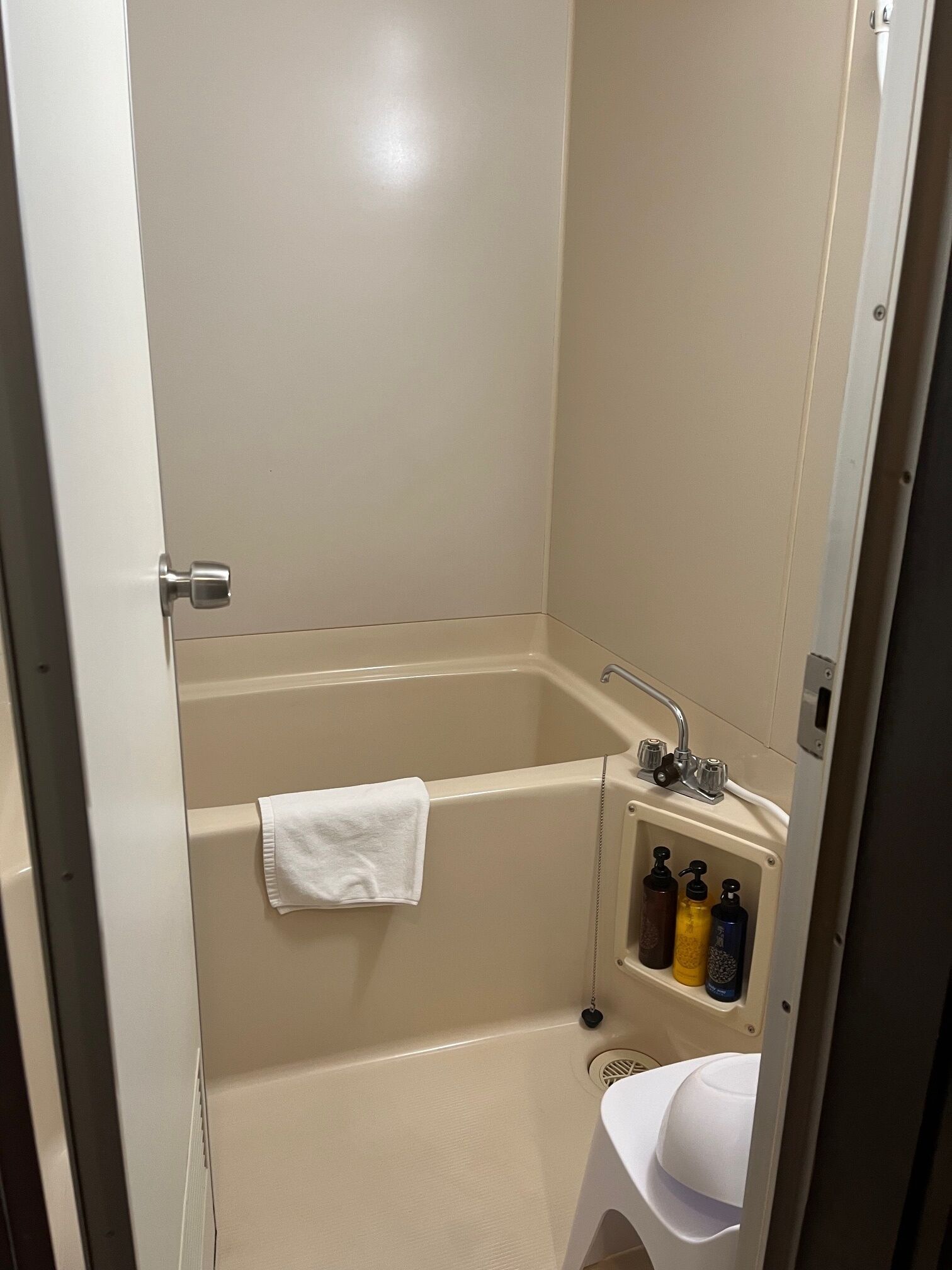 A small, enclosed bathroom with a bathtub, a white towel hanging on the wall, and a shelf with toiletries.