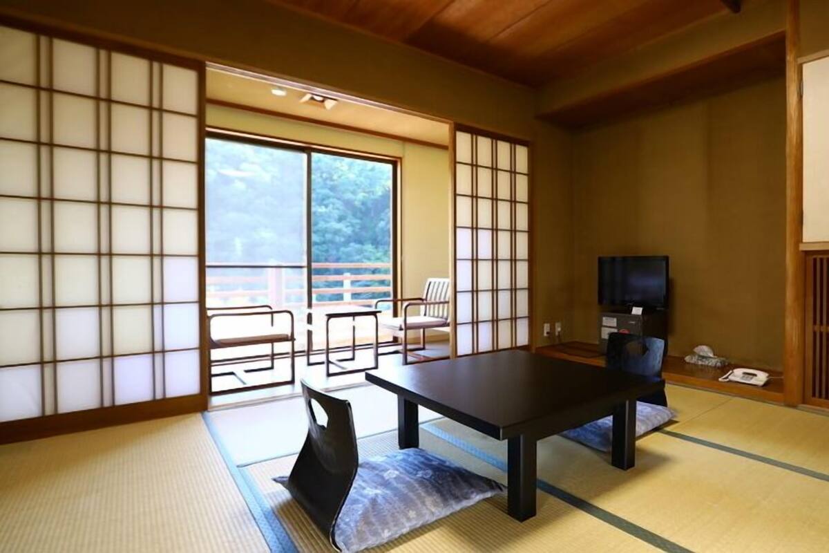 Une pièce de style japonais traditionnel, avec un sol recouvert de tatamis, une table basse, un téléviseur et des portes coulissantes donnant sur un espace extérieur.