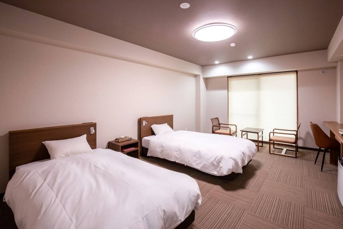 Une chambre d’hôtel avec deux lits, un bureau, une chaise et une grande fenêtre.